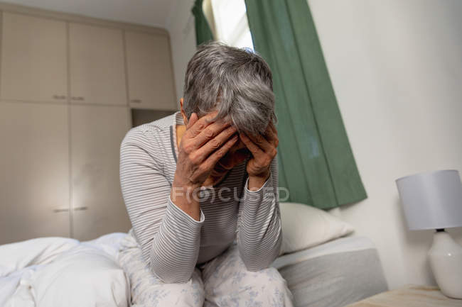 Front view close up of a mature Caucasian woman with short grey hair sitting on the side of her bed at home with her head in her hands — Stock Photo