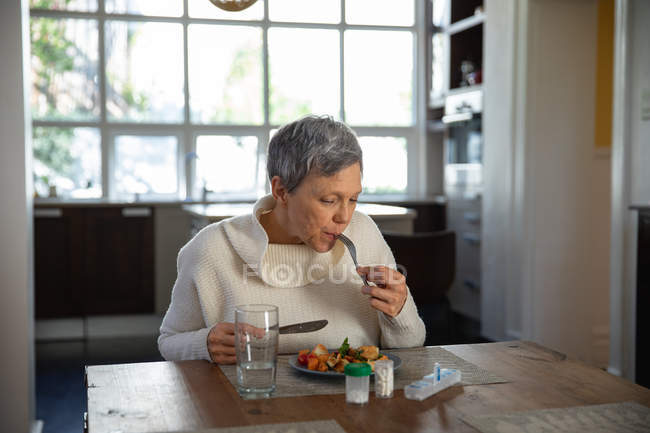 Вид на літні кавказькі жінки сидять і їдять на своєму обідньому столі, з склянку води, пляшки таблеток і коробку таблеток на стіл перед нею — стокове фото