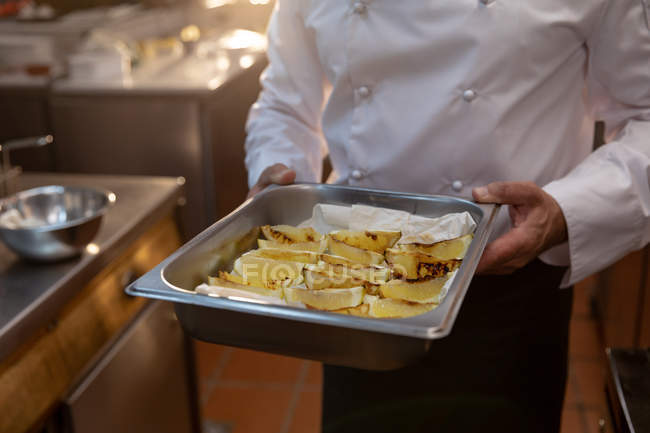 Vista frontal sección central de un chef masculino sosteniendo una bandeja para hornear de metal de limones cocidos en una cocina de restaurante - foto de stock
