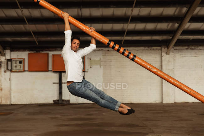Vista lateral de um jovem dançarino de balé segurando um poste estrutural e dançando com os pés fora do chão em uma sala vazia em um armazém abandonado — Fotografia de Stock