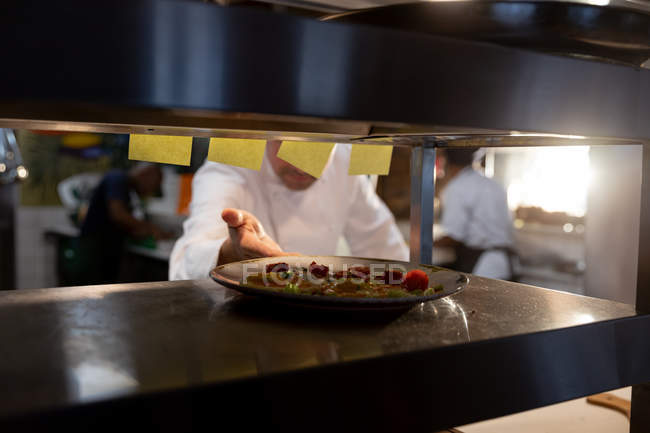 Front view close up of male chef reaching out for a plate of prepared food at the order station in a restaurant kitchen, seen through shelves — Stock Photo
