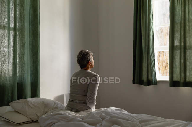 Заднього виду зрілої кавказької жінки з коротким сивим волоссям сидить на боці її ліжка в будинку, дивлячись з вікна — стокове фото