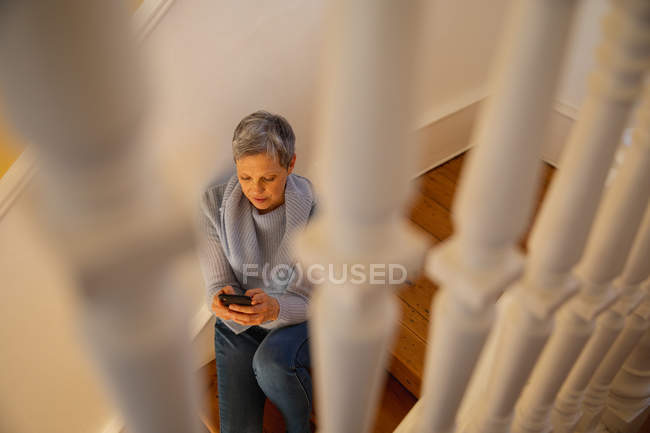 Elevated front view of a mature Caucasian woman with short grey hair sitting on her stairs at home using a smartphone — Stock Photo