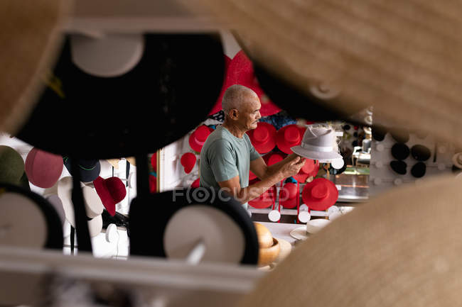 Вид збоку на старшого змішаного гоночного чоловіка, який сидить і оглядає готовий капелюх, оточений капелюхами на виставці в виставковому залі на капелюшній фабриці, видно через виставкову підставку — стокове фото