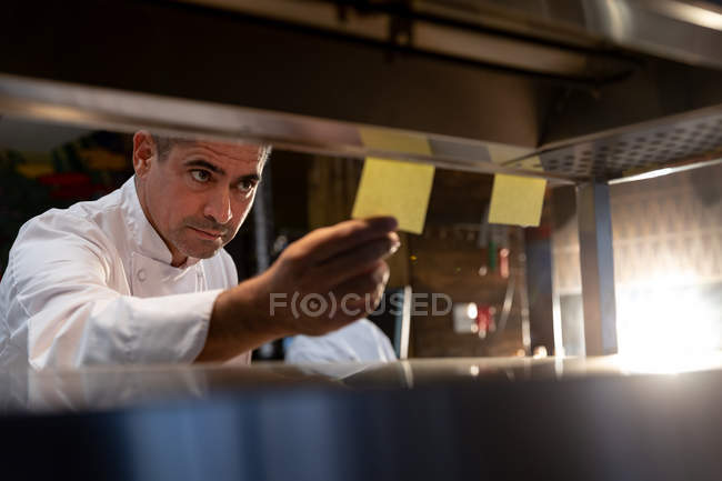 Nahaufnahme eines kaukasischen Küchenchefs mittleren Alters, der an der Bestellstation in einer Restaurantküche Bestellungen überprüft, durch die Regale gesehen — Stockfoto