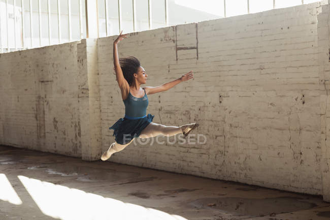 Вид збоку молодої змішаної раси танцівниця балету стрибає в повітрі з піднятими руками під час танців у порожній кімнаті на покинутому складі — стокове фото