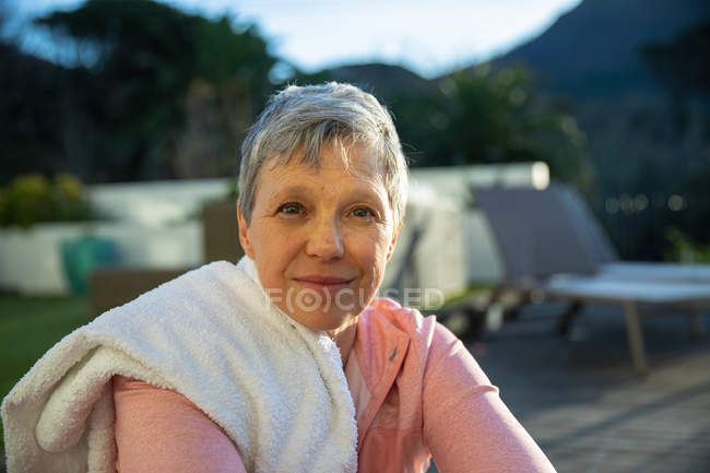 Porträt einer reifen kaukasischen Frau mit kurzen grauen Haaren, die nach dem Sport in ihrem Garten sitzt und leicht lächelnd in die Kamera blickt, mit einem Handtuch auf der Schulter — Stockfoto