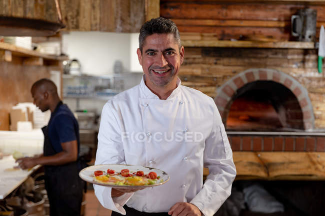 Retrato de cerca de un sonriente chef caucásico de mediana edad sosteniendo un plato de comida preparada en una cocina de restaurante, mientras que un miembro masculino del personal de cocina trabaja en el fondo - foto de stock
