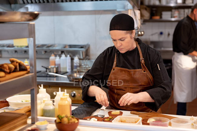 Front view close up of a young Caucasian female chef preparing dishes in a busy restaurant kitchen with other kitchen staff working in the background — Stock Photo