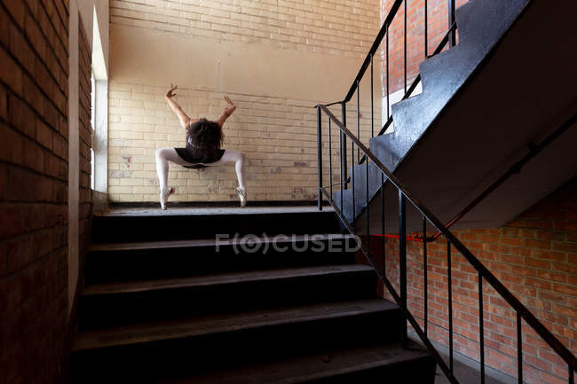 Vista frontal de una joven bailarina de ballet de raza mixta que sostiene una pose de baile en los dedos de los pies con los brazos levantados y la cabeza hacia abajo en una escalera que aterriza en un almacén abandonado - foto de stock