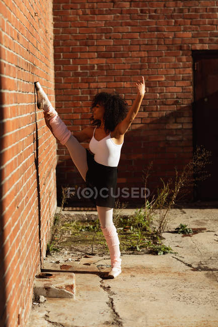 Side view close up of a young mixed race female ballet dancer standing on one leg on her toes holding her other leg up against a brick wall with one arm raised, on the rooftop of an urban building — Stock Photo