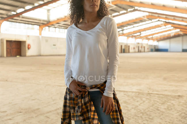 Vista frontal sección media de una joven mestiza con el pelo rizado y una camisa atada alrededor de su cintura de pie y mirando hacia otro lado en un almacén abandonado - foto de stock