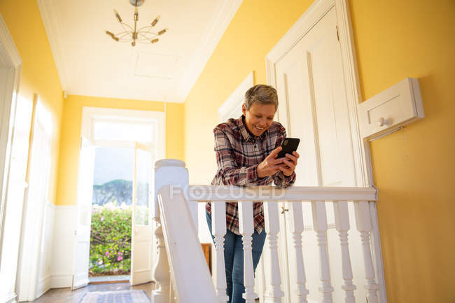 Вид спереди на улыбающуюся взрослую белую женщину с короткими седыми волосами, стоящую на посадочной площадке дома, опирающуюся на перила с помощью смартфона, окно с солнечным освещением на заднем плане с видом на сельскую местность — стоковое фото