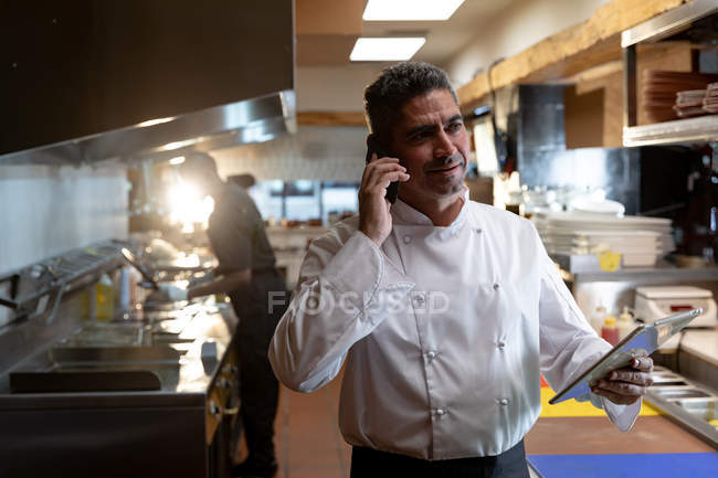 Vista frontal de cerca de un chef caucásico de mediana edad en el teléfono y sosteniendo una tableta en la cocina de un restaurante, un miembro del personal de la cocina que trabaja detrás de él - foto de stock