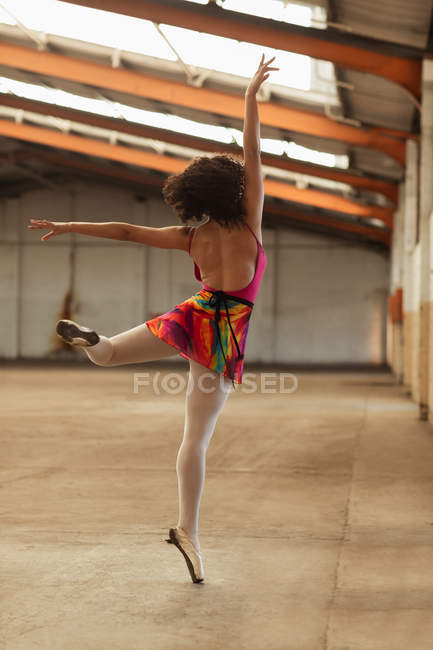 Vista posteriore da vicino di una giovane ballerina di danza mista in piedi su una gamba con le braccia tese mentre balla in una stanza vuota in un magazzino abbandonato — Foto stock