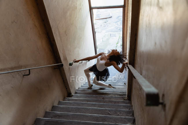 Vista laterale elevata di una giovane ballerina di danza mista che tiene i corrimano e balla in fondo a una scala in un magazzino abbandonato — Foto stock