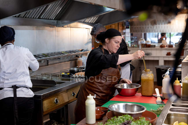 Seitenansicht einer jungen kaukasischen Köchin, die in einer Restaurantküche eine Soße aus einem Glas in eine Metallschüssel gießt, während andere Küchenmitarbeiter im Hintergrund arbeiten — Stockfoto