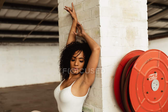 Seitenansicht einer jungen Balletttänzerin mit gemischter Rasse, die mit erhobenen Armen und dem Rücken gegen eine Säule in einem leeren Raum einer verlassenen Lagerhalle tanzt — Stockfoto