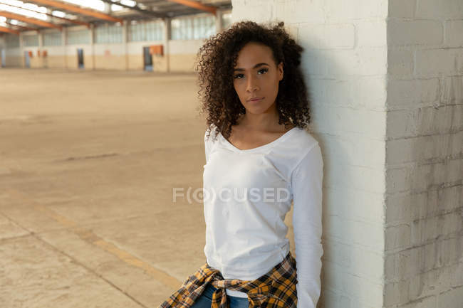 Front view close up of a young mixed race woman with shoulder length curly hair and a shirt tied around her waist leaning against a pillar and looking to camera in an abandoned warehouse — Stock Photo