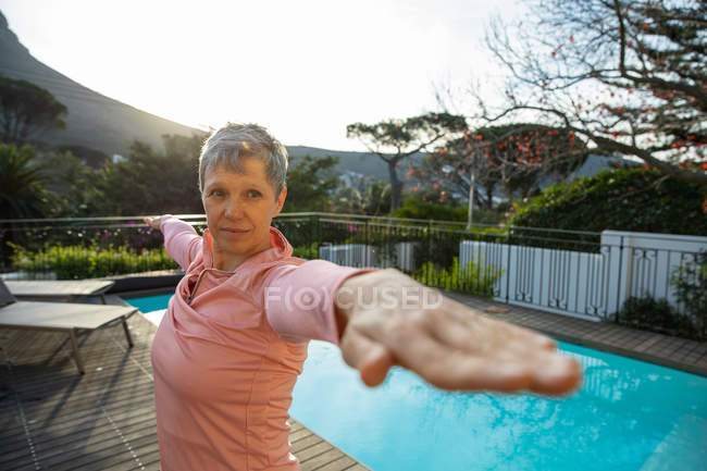 Vista frontal close-up de uma mulher branca madura com cabelos brancos curtos em pé em uma posição de ioga, exercitando-se junto à piscina em seu jardim, com uma vista rural no fundo — Fotografia de Stock