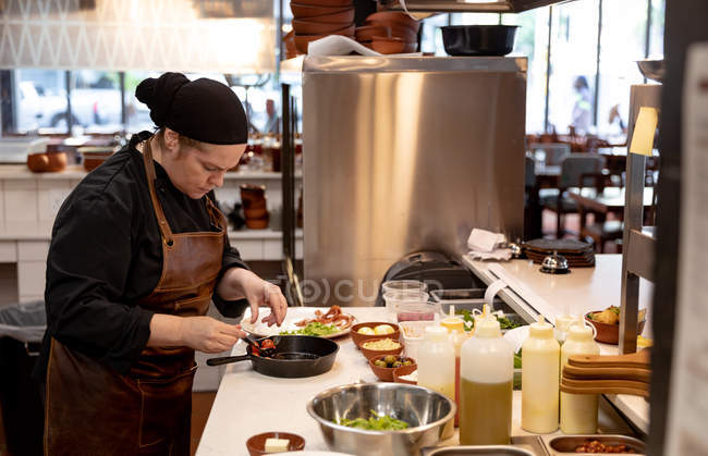 Vue de côté gros plan d'une jeune cuisinière caucasienne ajoutant des ingrédients à une casserole dans une cuisine de restaurant — Photo de stock