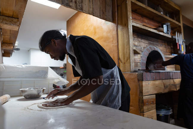 Vista lateral de cerca de un joven trabajador de cocina afroamericano que prepara masa para una base de pizza en una cocina de restaurante, mientras que otro miembro del personal de cocina trabaja en el horno de pizza en el fondo - foto de stock