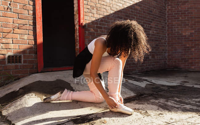 Vista lateral de cerca de una joven bailarina de ballet de raza mixta arrodillada y atando su zapato puntiagudo en la azotea de un almacén abandonado - foto de stock