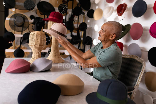 Вид збоку на старшого змішаного гоночного чоловіка, який сидить і оглядає готовий капелюх, оточений капелюхами на виставці в виставковому залі на капелюшній фабриці — стокове фото