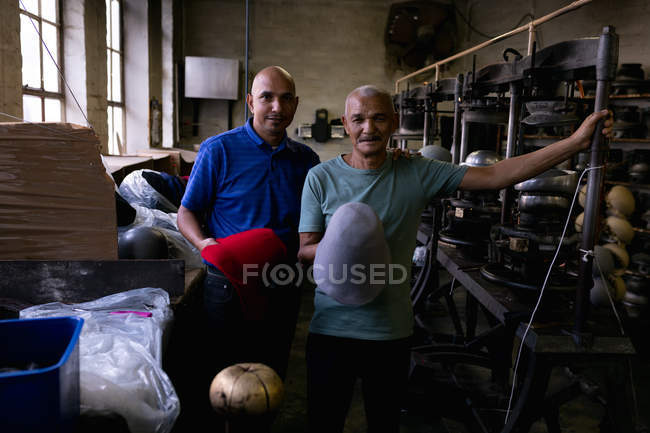 Portrait of a smiling senior and middle aged mixed race men holding the tops of two hats that have been shaped on equipment they stand beside in the workshop at a hat factory — Stock Photo