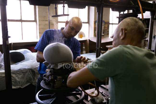Por encima de la vista del hombro de un senior y un mestizo de mediana edad trabajando juntos en una máquina humeante la parte superior de un sombrero para darle forma en el taller en una fábrica de sombreros - foto de stock