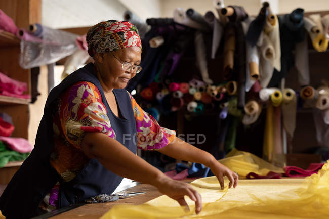 Vista laterale da vicino di una donna di razza mista di mezza età in piedi a un tavolo che lavora con tessuto giallo in una fabbrica di cappelli . — Foto stock