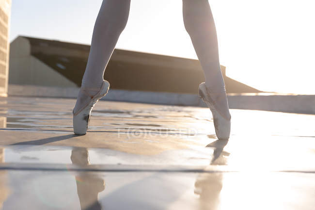 Vista frontal de la sección baja de la bailarina de ballet con medias y zapatos de punta, de pie sobre sus dedos de los pies en la azotea de un edificio urbano, retroiluminado por la luz del sol - foto de stock