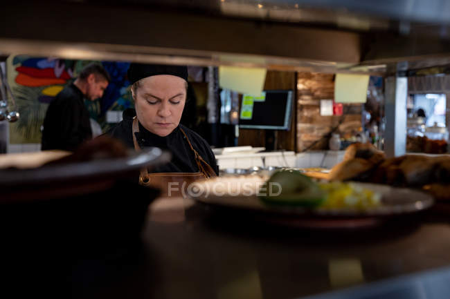 Front view close up of a young Caucasian female chef at work in a busy restaurant kitchen, seen through shelves, with other kitchen staff working in the background — Stock Photo