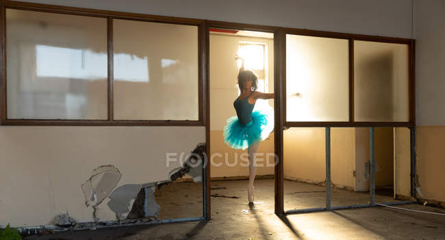 Vista frontal de una joven bailarina de ballet de raza mixta con un tutú azul y zapatos puntiagudos bailando sobre una pierna en una puerta en un edificio abandonado del almacén, retroiluminado por la luz del sol - foto de stock