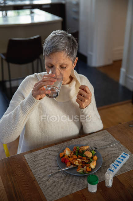 Vista frontal elevada de uma mulher branca madura sentada em sua mesa de jantar bebendo um copo de água, com comida em um prato e garrafas de comprimidos e pílulas na mesa na frente dela — Fotografia de Stock