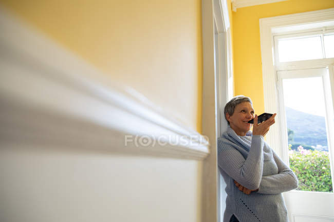 Vista lateral de uma mulher branca madura com cabelos brancos curtos apoiados em uma parede em casa falando em um smartphone que ela está segurando na frente da boca e sorrindo, com uma cena rural visível pela janela no fundo — Fotografia de Stock
