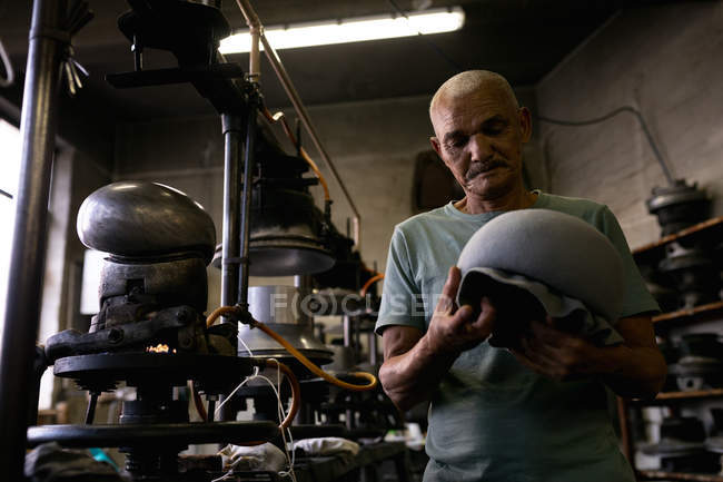 Vista frontal de cerca de un hombre de raza mixta senior sosteniendo e inspeccionando la parte superior de un sombrero que ha sido moldeado en una pieza de equipo en el taller en una fábrica de sombreros - foto de stock