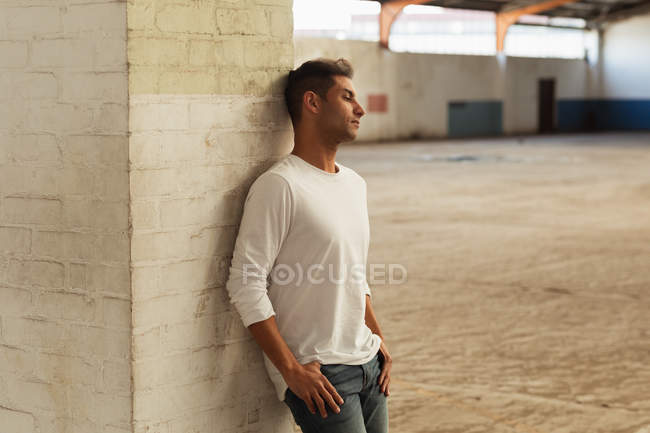 Seitenansicht eines jungen Mannes, der sich mit den Daumen in den Hosentaschen an eine Säule lehnt und in einem leeren Raum einer verlassenen Lagerhalle wegblickt — Stockfoto