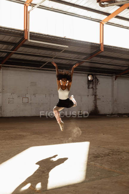 Front view of a young mixed race female ballet dancer wearing pointe shoes jumping in the air in shaft of sunlight with arms raised while dancing in an empty room at an abandoned warehouse — Stock Photo