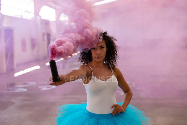 Vista frontal de cerca de una joven bailarina de ballet de raza mixta con un tutú azul, sosteniendo una granada de humo rosa y mirando a la cámara en una habitación vacía en un almacén abandonado - foto de stock