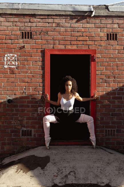 Vista frontal close-up de uma jovem mestiça dançarina de balé feminina segurando uma pose de dança em uma porta em uma parede de tijolo e olhando para a câmera, no telhado de um edifício urbano — Fotografia de Stock