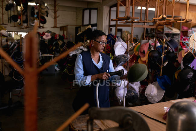 Vista frontal de una mujer de raza mixta de mediana edad que opera una máquina para limpiar al vapor un sombrero que sostiene, de pie en una mesa en el taller en una fábrica de sombreros, otros sombreros visibles en el fondo - foto de stock