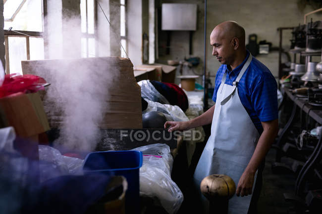 Side view of a middle aged mixed race man wearing an apron operating a piece of equipment in the workshop at a hat factory, with steam and materials in the foreground — Stock Photo