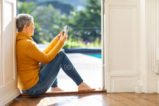 Vista laterale da vicino di una donna caucasica matura seduta sulla porta del giardino di casa con uno smartphone, con alberi illuminati dal sole sullo sfondo — Foto stock