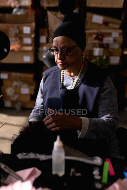 Vista frontal close-up de uma mulher de meia idade mista segurando e inspecionando um pequeno chapéu na oficina em uma fábrica de chapéus, com caixas de materiais no fundo — Fotografia de Stock