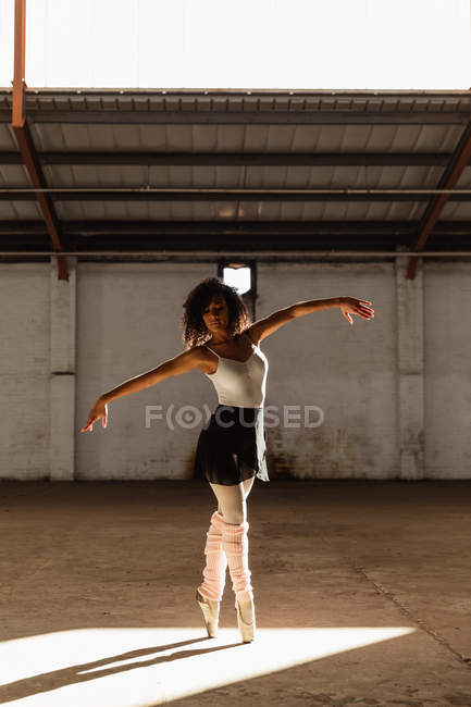 Vista frontal de una joven bailarina de ballet de raza mixta con zapatos puntiagudos de pie sobre sus dedos de los pies en el eje de la luz solar con los brazos extendidos mientras baila en una habitación vacía en un almacén abandonado - foto de stock