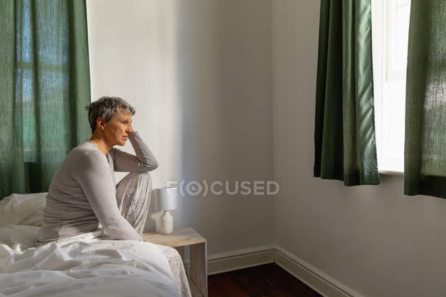Vista lateral de una mujer caucásica madura con el pelo gris corto sentado en el lado de su cama en casa apoyado en su rodilla levantada, mirando hacia otro lado - foto de stock