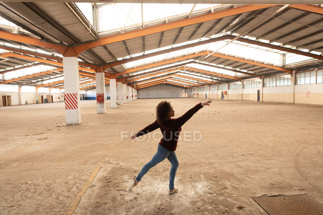 Side view of a young mixed race female ballet dancer wearing jeans and pointe shoes dancing with arms outstretched in an abandoned warehouse — Stock Photo