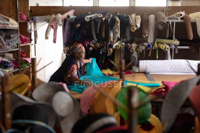 Vista elevada de una mujer mestiza de mediana edad parada en una mesa trabajando con tela azul en una fábrica de sombreros . - foto de stock