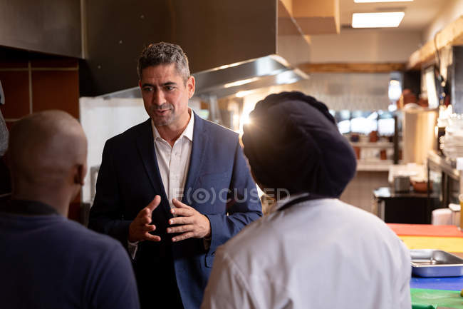 Vista frontal de cerca de un gerente de restaurante caucásico de mediana edad hablando con dos miembros del personal de la cocina, visto desde la parte posterior, en una cocina de restaurante - foto de stock